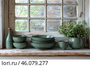 A cozy still life on a windowsill with vintage elements. On an old wooden window frame, slightly peeling from time, there is greenish ceramic tableware with a craquelure effect. The set includes plates, bowls, cups and a bottle with a cork. On the right is a vase with white wild flowers, giving the composition naturalness and freshness. Blurred outlines of a brick wall and greenery are visible outside the window. The atmosphere of the image conveys a sense of coziness, rustic style and warm home traditions. Стоковое AI-изображение, AI-художник Анна Кривицкая / Фотобанк Лори
