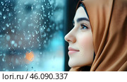 A young woman with a hijab contemplates life as raindrops cascade down the window beside her. Стоковое AI-изображение, AI-художник Елена Старчак / Фотобанк Лори