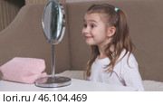Fashion little girl in a white T-shirt is sitting at the dressing table in front of the mirror, smiling and winking her eyes. Advertising of children's cosmetics concept. Footage for kids blog. Стоковое видео, видеограф Ирина Михайличенко / Фотобанк Лори