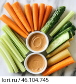 A healthy snack consisting of freshly sliced ​​vegetables neatly arranged on a marble surface. In the center are two small white cups filled with what looks like a nut or tahini dressing. Around them are crunchy vegetables: carrots, celery, fresh cucumber, and perhaps a pickled rhubarb stalk or another variety of celery. The color palette of the image is rich in greens, oranges, and creams, creating a delicious and aesthetically pleasing look. Стоковое AI-изображение, AI-художник Анна Кривицкая / Фотобанк Лори