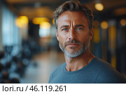 A masculine middle-aged man with grey stubble and thick hair combed back. His eyes are blue and his expression is confident and calm. He is wearing a grey sports T-shirt. The background is blurred, but it is clear that he is in a gym, where you can see the sports equipment, dumbbells and soft lighting.The overall style of the photo is a modern and stylish portrait, conveying strength, maturity and confidence. The man looks well-groomed, perhaps plays sports and takes care of himself. The atmosphere of the photo creates a sense of calm determination and inner strength. Стоковое AI-изображение, AI-художник Анна Кривицкая / Фотобанк Лори