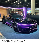A futuristic sports car concept exhibited at a car show. The car is painted in deep black with neon purple highlights that emphasize its aerodynamic silhouette and aggressive style.The front of the car is distinguished by a mesh-shaped radiator grille, narrow LED headlights and a low profile body. The car sits on black multi-spoke wheels, and its reflective surface catches the light reflections of the showroom.The background is a car show, with a modern design in purple and wood tones, digital displays and visitors. LED elements on the car create a cyberpunk aesthetic. Стоковое AI-изображение, AI-художник Анна Кривицкая / Фотобанк Лори