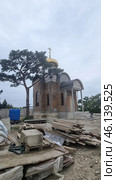 An Orthodox church under construction with a golden dome and an Orthodox cross on top. The walls of the building are made of red brick, in some places not yet covered with a finishing layer. The architectural style is reminiscent of traditional Russian churches, with characteristic semicircular arches above the windows and entrance. Стоковое фото, фотограф Анна Кривицкая / Фотобанк Лори
