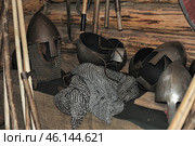 Elements of medieval military equipment, including metal helmets, chain mail and weapons. One of the helmets has a visor and a chain mail aventail, which is typical for medieval knights or warriors of Ancient Rus'. The other helmets have an open design and leather straps for fixation. In the foreground lies a chain mail shirt made of many metal rings. In the background, you can see various axes, clubs and other types of bladed weapons hanging on a wall made of logs. The whole scene resembles a military arsenal or a museum exhibition associated with historical reconstruction. Стоковое фото, фотограф Анна Кривицкая / Фотобанк Лори