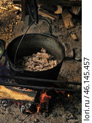 A camp cast iron cauldron hanging over an open fire in which meat and onions are being cooked. Pieces of meat are fried in boiling fat, and under the cauldron a fire burns, fueled by wooden logs. The atmosphere is reminiscent of a traditional field kitchen, perhaps of Vikings, nomads or hunters.The background of the scene is decorated with sandy soil, scattered firewood and traces of a fire pit. Such cooking is typical for camping conditions, fishing camps or historical reconstructions related to the life of ancient peoples. Стоковое фото, фотограф Анна Кривицкая / Фотобанк Лори