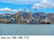 Novorossiysk Commercial Sea Port, one of the largest ports in Russia, located on the Black Sea coast. The frame shows cargo ships, port cranes, container yards and part of the industrial infrastructure. In the background are the mountain ranges surrounding the city.Numerous blue and yellow gantry cranes used for loading and unloading ships.Cargo containers stored in the port area.Commercial ships at the berths undergoing cargo operations.Tugboats used to maneuver large ships.Mountains, characteristic of the Novorossiysk landscape.Bright blue sky with clouds, creating a contrast with the industrial landscape. (2023 год). Редакционное фото, фотограф Анна Кривицкая / Фотобанк Лори