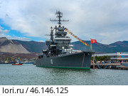 The cruiser Mikhail Kutuzov, permanently moored at the Novorossiysk embankment. This is one of the main museum ships of Russia, which is part of the Central Naval Museum.The cruiser Mikhail Kutuzov is a massive warship with the number "105" on board.It is decorated with signal flags, creating a festive atmosphere.In the background, the port of Novorossiysk, the mountains of the Markotkhsky Range and the berthing facilities are visible.The red naval flag with a white star flutters at the stern.To the right of the ship is the marine terminal, where people and city infrastructure are visible.In the distance, a small tug or boat moves along the water. (2023 год). Редакционное фото, фотограф Анна Кривицкая / Фотобанк Лори