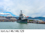 The cruiser Mikhail Kutuzov, permanently moored at the Novorossiysk embankment. This is one of the main museum ships of Russia, which is part of the Central Naval Museum.The cruiser Mikhail Kutuzov is a massive warship with the number "105" on board.It is decorated with signal flags, creating a festive atmosphere.In the background, the port of Novorossiysk, the mountains of the Markotkhsky Range and the berthing facilities are visible.The red naval flag with a white star flutters at the stern.To the right of the ship is the marine terminal, where people and city infrastructure are visible.In the distance, a small tug or boat moves along the water. (2023 год). Редакционное фото, фотограф Анна Кривицкая / Фотобанк Лори