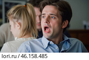Shocked young man with open mouth wide eyes looking surprised wearing blue shirt sitting indoors with people in background feeling unexpected fear or surprise. Стоковое AI-изображение, AI-художник Павел Муравьев / Фотобанк Лори