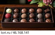 A box with exquisite chocolates, neatly placed in individual compartments. Each candy differs in shape, texture and decoration: there is milk, dark and white chocolate, decorations made of cocoa powder, nut crumbs, caramel and colored glaze. One of the candies is covered in red sugar, standing out from the rest. Flowers are visible in the background, which adds aesthetics to the image and the atmosphere of a gift or a special occasion. The overall style conveys luxury, taste and pleasure from chocolate. Стоковое AI-изображение, AI-художник Анна Кривицкая / Фотобанк Лори