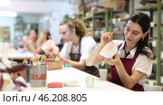 Smiling young girl in apron painting on handmade ceramic bowl with brush in pottery workshop. Стоковое видео, видеограф Яков Филимонов / Фотобанк Лори