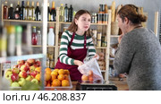 Friendly smiling young saleswoman in striped jumper and apron chatting warmly with male customer while weighing fresh tangerines in cozy grocery store. Стоковое видео, видеограф Яков Филимонов / Фотобанк Лори