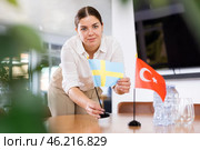 Positive young woman putting flag of Sweden on the table with flag of Turkey in conference room. Стоковое фото, фотограф Яков Филимонов / Фотобанк Лори
