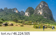 Picturesque karst mountains against a clear blue sky. There is a round hole at the top. Green vegetation on steep slopes, in the valley. Boulders in the meadow. People are walking around the clearing. Стоковое фото, фотограф Вера Ивановна Кубарева / Фотобанк Лори