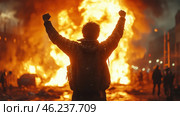 A dramatic scene of rioting or rebellion. In the center stands a man in dark clothing with his fists raised in triumph, determination, or protest. Behind him, a huge fire rages across the street, perhaps from burning cars or buildings. In the distance, dark silhouettes of other people can be seen, who may be rioters or bystanders. The atmosphere is unsettling, filled with chaos and dramatic lighting from the fire. Стоковое AI-изображение, AI-художник Анна Кривицкая / Фотобанк Лори