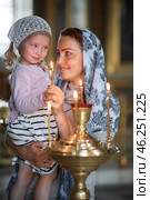 Russian beautiful woman in a scarf and with red hair holding a little girl and lights a candle in front of an icon in the Russian Orthodox Church. Стоковое фото, фотограф Арзюков Вадим Николаевич / Фотобанк Лори