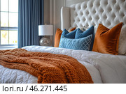 A cozy bedroom with an elegant interior. In the center of the frame is a large bed with a white quilted bedspread, decorated with decorative pillows in shades of orange and blue. The pillows are made of velvety fabrics, which gives the atmosphere luxury and comfort. On top of the bed lies a warm, large-knit plaid in terracotta, which complements the color scheme of the room. At the head of the bed is a soft white headboard with a capitonné. In the background, you can see a window with long curtains and a stylish bedside lamp with a round glass base. Стоковое AI-изображение, AI-художник Анна Кривицкая / Фотобанк Лори
