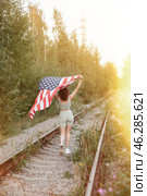 A woman runs joyfully along a railway track, waving an American flag. The warm sunlight filters through the trees, creating a serene and festive atmosphere as she embraces her spirit of freedom. Стоковое фото, фотограф Aleksandr Golubev / Фотобанк Лори