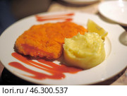 A dish consisting of a breaded cutlet, fried until crispy, with a side of mashed potatoes. The cutlet is covered in bright crispy breading and topped with ketchup, which is also beautifully spread on the plate. Next to the puree is a slice of lemon, which can serve as an addition to the meat. The dish is served on a white plate in a cozy restaurant setting. Стоковое фото, фотограф Анна Кривицкая / Фотобанк Лори