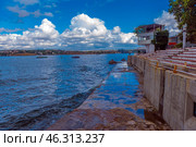 A picturesque view of Sevastopol Bay with a concrete embankment and the steps of an amphitheater facing the sea. Along the shore there is a white building with a terrace, and in the distance you can see ships and cranes of shipyards. The weather is sunny but cloudy, creating a striking contrast between the blue sky, sea and architecture. On the horizon you can see the cityscape of Sevastopol with its historical and industrial sites. (2021 год). Редакционное фото, фотограф Анна Кривицкая / Фотобанк Лори