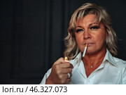 A middle-aged woman sits indoors, focused and contemplative. She lights a cigarette, dressed in a crisp white shirt. The atmosphere is intimate, capturing a moment of solitude and thoughtfulness. Стоковое фото, фотограф Aleksandr Golubev / Фотобанк Лори