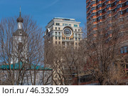 Architectural contrast between historical and modern buildings.On the left is the Church of the Resurrection of the Word in Barashi, known for its old architectural style. Its white walls and dark dome with a cross stand out against the blue sky.The center is dominated by a skyscraper with a huge clock - this is the famous House with a Clock on Chistoprudny Boulevard. It is built in the neoclassical style and is known for its luxurious facade with columns and a dome. The clock gives the building a unique look.On the right, you can see a modern apartment building with red-brown panels, which reflects the more utilitarian style of Moscow architecture of the late 20th - early 21st centuries.The trees without leaves indicate that the photo was taken in late autumn or early spring. Стоковое фото, фотограф Анна Кривицкая / Фотобанк Лори
