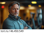 A mature man with graying hair and beard, wearing a gray sports hoodie with a zipper. He is standing in a gym or fitness center, arms crossed over his chest, with a confident and serious expression on his face. The background is blurred, but you can see the exercise machines and soft lighting, creating a warm but professional atmosphere. The man's gaze is focused, which may indicate his experience and self-confidence. Стоковое AI-изображение, AI-художник Анна Кривицкая / Фотобанк Лори