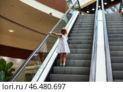 A young child dressed in a white outfit curiously stands on an escalator, reaching out towards the rail. Surrounded by a contemporary shopping center, the scene conveys a sense of wonder. Стоковое фото, фотограф Aleksandr Golubev / Фотобанк Лори