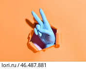 Рука в медицинской перчатке показывает победный жест сквозь дыру в бумаге на цветном фоне. Стоковое фото, фотограф Pixel_shot / Фотобанк Лори