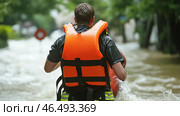 Rescuer wearing life vest walking through floodwaters during rescue operation. Стоковое AI-изображение, AI-художник Юрий Поздников / Фотобанк Лори
