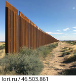 A massive metal border wall stretching into the distance. It is made of rusty steel panels and rises above a desert landscape. To the right of the wall there is a dusty road overgrown with bushes and low vegetation. The horizon is clear, the sky is blue with small clouds, and in the distance you can see the silhouettes of mountains. The scene creates an atmosphere of isolation, border and division of territories. Стоковое AI-изображение, AI-художник Анна Кривицкая / Фотобанк Лори