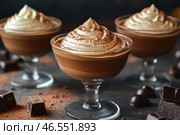 Three servings of chocolate mousse in elegant glass goblets. The dessert is topped with a thick cap of whipped cream, generously sprinkled with cocoa powder. In the foreground, you can see pieces of dark chocolate and scattered cocoa powder, which adds depth to the composition and emphasizes the chocolate theme. The background is darkened, creating a cozy and atmospheric setting, focusing attention on the texture and richness of the dessert. Overall, the image conveys a sense of exquisite taste and gastronomic pleasure. Стоковое AI-изображение, AI-художник Анна Кривицкая / Фотобанк Лори