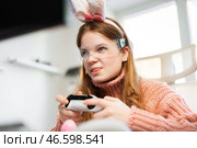 Passionate gamer girl in bunny ears and pink sweater competes in Easter-themed game. Holding controller, she focuses on screen, enjoying festive cyber gaming session. Стоковое фото, фотограф Енваев Андрей / Фотобанк Лори