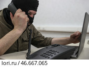 Мошенник в черной балаклаве у ноутбука с телефонной трубкой в руке. Стоковое фото, фотограф Сергей Булкин/NEWS.ru / ТАСС / Фотобанк Лори