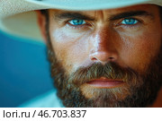 A close-up portrait of a man with a brutal appearance. He has bright blue eyes, a thick beard and tanned skin with a pronounced texture. He wears a light hat, possibly a cowboy hat, which gives the image a characteristic western atmosphere. The look is penetrating, serious, with a hint of mystery. The background is blurred, made in cold shades, creating a contrast with the warm tone of the skin and hair. The image evokes associations with masculinity, strength and the spirit of adventure. Стоковое AI-изображение, AI-художник Анна Кривицкая / Фотобанк Лори
