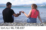 Man and woman clinging glasses of red wine while sitting on the pebble beach by the sea. Стоковое фото, фотограф David Kashakhi / Фотобанк Лори
