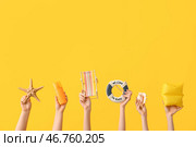 Женские руки с солнцезащитным кремом, морская звезда, миниатюрный шезлонг, декоративный спасательный круг, ракушка и надувной нарукавник на желтом фоне. Концепция путешествий. Стоковое фото, фотограф Pixel_shot / Фотобанк Лори