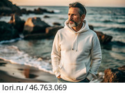 A handsome middle-aged man with a beard wearing a white hoodie on the beach. Template layout for hoodie design, print area layout for logo or design. Стоковое AI-изображение, AI-художник Photojuli / Фотобанк Лори