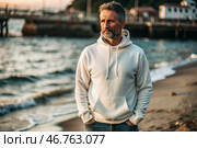 A handsome middle-aged man with a beard wearing a white hoodie on the beach. Template layout for hoodie design, print area layout for logo or design. Стоковое AI-изображение, AI-художник Photojuli / Фотобанк Лори