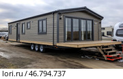 A modern mobile home on wheels with an elegant design. The house is made in gray and wooden shades, equipped with large panoramic windows and a spacious terrace with a wooden staircase. It stands on an open area among other mobile homes and caravans, which indicates the place of sale or temporary parking. The house looks spacious, comfortable for living and adapted for traveling or autonomous living. Стоковое AI-изображение, AI-художник Анна Кривицкая / Фотобанк Лори