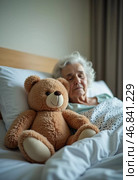 In a quiet hospital room, an elderly woman rests peacefully in bed, finding solace in the soft embrace of a teddy bear by her side, symbolizing companionship through hardship. Стоковое AI-изображение, AI-художник Юлия Петровская / Фотобанк Лори
