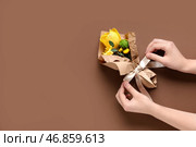 Женские руки с мини-букетом красивых желтых весенних цветов в упаковочной бумаге на коричневом фоне. Международный женский день. Стоковое фото, фотограф Pixel_shot / Фотобанк Лори