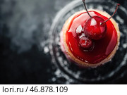 An exquisite dessert with a bright red glaze that gives it an appetizing shine. On top of the dessert are two cherries with thin branches, covered with powdered sugar. The base of the dessert is a delicate sponge cake or shortcrust pastry that contrasts with the rich glossy glaze. The background of the image is dark and slightly blurred, which emphasizes the brightness of the dessert and its attractive appearance. Стоковое AI-изображение, AI-художник Анна Кривицкая / Фотобанк Лори