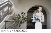 Thoughtful young bride in gorgeous long wedding dress holding bouquet and standing near the house with stairs. Стоковое фото, фотограф David Kashakhi / Фотобанк Лори