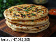 A stack of flavorful, crispy flatbreads that have been cooked on a griddle or grill. They are golden brown in color with delicious toasted spots. The flatbreads are garnished with fresh herbs, perhaps cilantro or parsley, adding a fresh flavor. The dish is served on a wooden board, with sauces or gravies slightly blurred in the background, hinting at the flatbread's oriental or Indian origins. Стоковое AI-изображение, AI-художник Анна Кривицкая / Фотобанк Лори