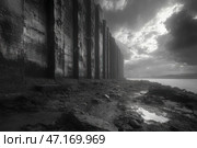 A dark industrial scene with a massive concrete wall stretching into the distance. Tall, rough columns covered in traces of time and erosion create a sense of abandonment and a post-apocalyptic atmosphere. The ground in front of the wall is littered with rocks, puddles and mud, adding to the harshness of the scene. The sky is covered with heavy clouds, through which a little light breaks through, creating dramatic lighting. On the horizon, beyond the water surface, the outlines of a factory or industrial complex are visible, complementing the industrial aesthetic of the frame. The black and white palette emphasizes the atmosphere of desolation, loneliness and time frozen in these concrete structures. Стоковое AI-изображение, AI-художник Анна Кривицкая / Фотобанк Лори