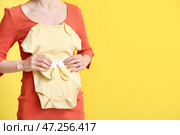 Молодая беременная женщина в детском боди и носках на желтом фоне. Стоковое фото, фотограф Pixel_shot / Фотобанк Лори