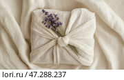 Creative gift wrapping technique using fabric and lavender for a personal touch at home. Красиво упакованный подарок демонстрирует элегантную ткань с веточкой лаванды, повязанной сверху. Этот декор придает подарку индивидуальность и продуманность. Стоковое AI-изображение, AI-художник Katsiaryna Repich / Фотобанк Лори