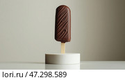 Chocolate coated popsicle stands on a white pedestal against a beige background creating a simple yet elegant display. Шоколадное мороженое стоит на белом пьедестале на бежевом фоне, создавая простой, но элегантный образ. Стоковое AI-изображение, AI-художник Katsiaryna Repich / Фотобанк Лори