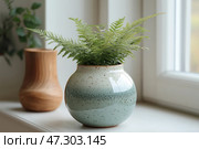 Interior still life with a ceramic pot with a green fern growing in it. The pot has a rounded shape and is covered with a glaze with a gradient from white to soft green with small specks, creating a handmade effect. The plant looks fresh, its delicate leaves spread out beautifully to the sides. A wooden vessel and blurred vegetation are visible in the background, and the composition itself is located on a windowsill near the window, which gives the scene soft natural lighting. The overall atmosphere conveys a sense of coziness, minimalism and closeness to nature. Стоковое AI-изображение, AI-художник Анна Кривицкая / Фотобанк Лори